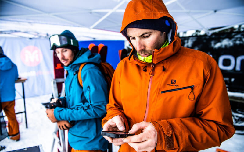 Ski test event in the French Alps – Man in an orange Salomon jacket using his phone in a snowy setting, with another skier in the background. A winter gear testing experience for ski enthusiasts.